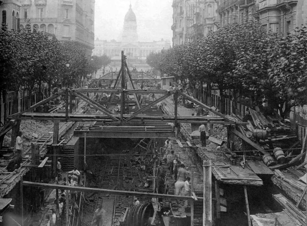Panoramic view of construction workers excavating for Buenos Aires's first subway line (Line A) below Avenida de Mayo in 1912, with surrounding buildings and early 20th-century urban scene.