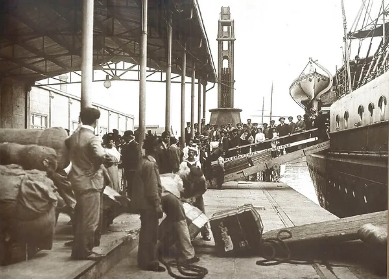 A large group of European immigrants with their belongings disembarking from a transatlantic ship at the bustling Port of Buenos Aires, with a tall tower in the background, circa 1900-1910.